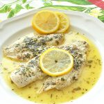 2 pieces of white fish served on a white plate smothered in lemon sauce and top with a slice of lemon.