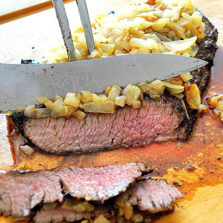 A grilled steak with caramelized onions on top is sliced on a wood carving boardl/