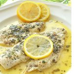 White fish cooked and served in a white place smothered in lemon sauce with sliced lemon on top.