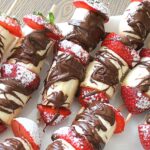 A white platter filled with skewers filled with sliced banana and strawberries with chocolate.