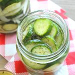 An open jar filled with pickles and dill set on a red and white checkered table cloth.