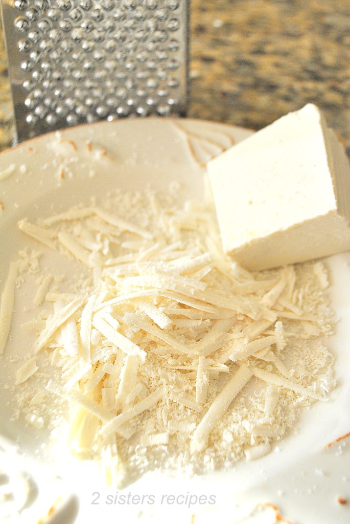 A wedge of Ricotta Salata cheese is grated into a plate.  by 2sistersrecipes.com