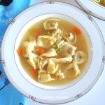 A white plate filled with tortellini in a broth with chopped carrots and celery.
