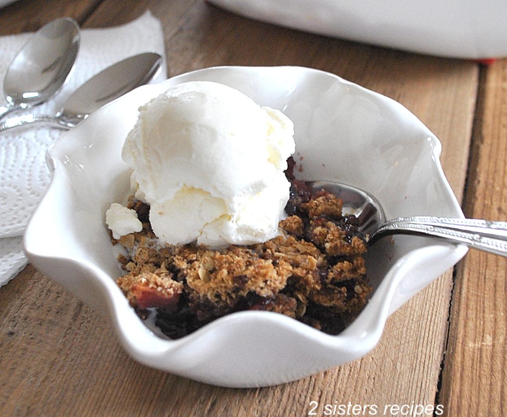 A white bowl filled with an apple cherry crisp with a scoop of vanilla ice cream on top.