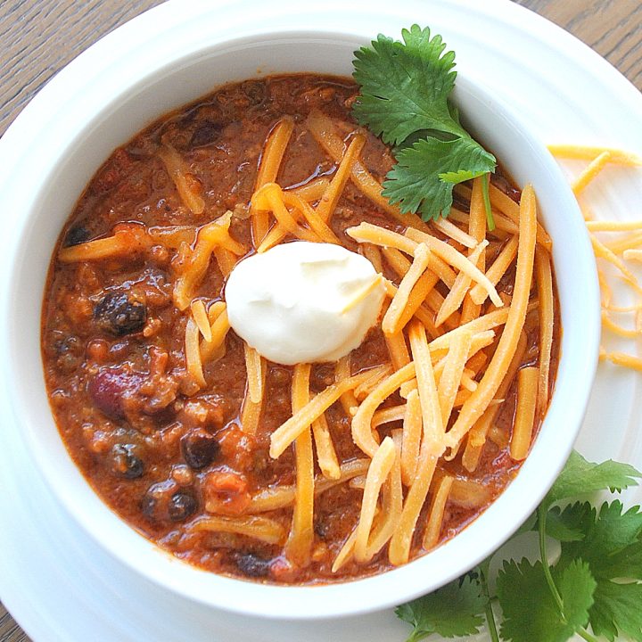 A white bowl of beef and beans with shredded cheese and a dollop of sour cream on top.
