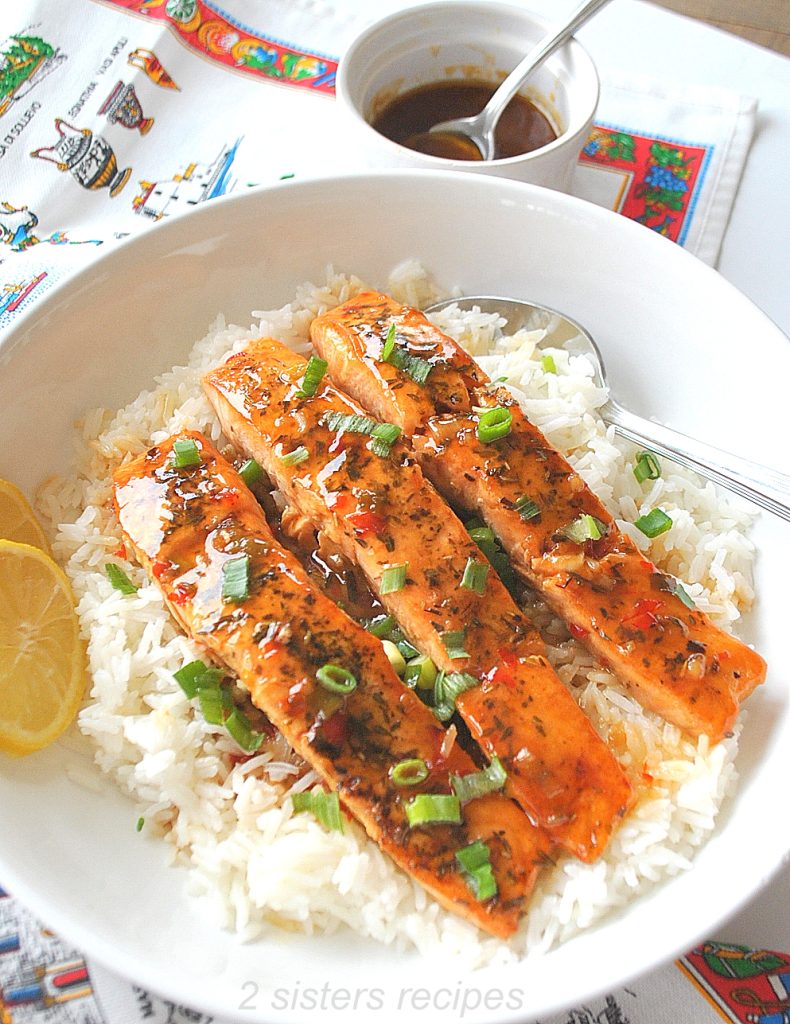 3 pieces of cooked salmon on top of steamed white rice with chopped green onions over them, on a white plate.