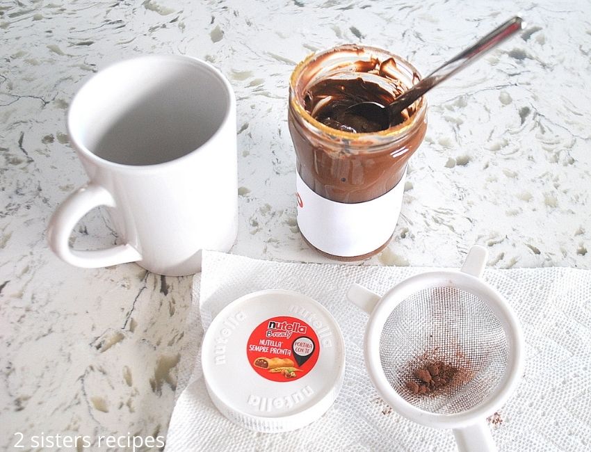 an opened jar of Nutella spread beside a white coffee mug. by 2sistersrecipes.com
