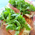 Two French bread slices served on a wood board topped with cheese, prosciutto and arugula.