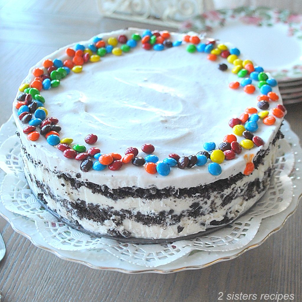 An layered ice cream cake topped with whipped cream and mini colorful candies served on a cake plate.