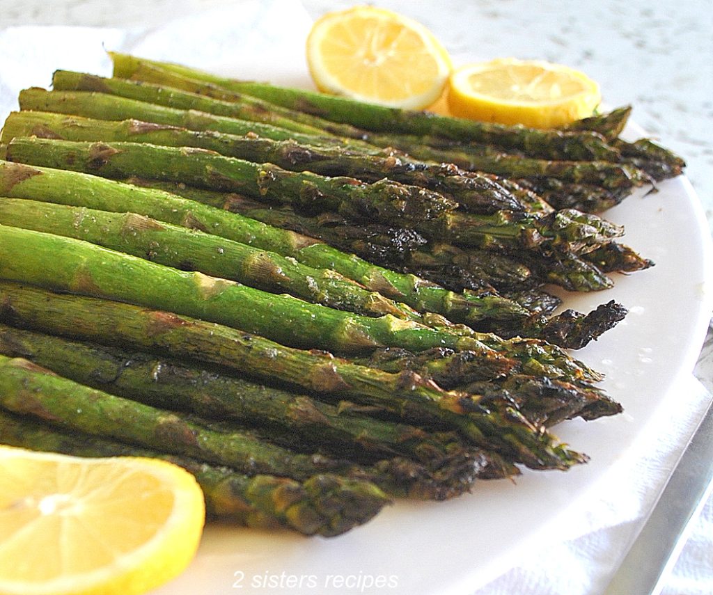 A white plate with grilled asparagus and lemon wedges. by 2sistersrecipes.com