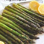 A bunch of asparagus grilled and served on a white platter with lemon wedges on the side.