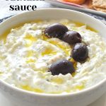 Authentic Tzatziki Sauce is served in a white bowl with a drizzle of olive oil and a few olives on top.