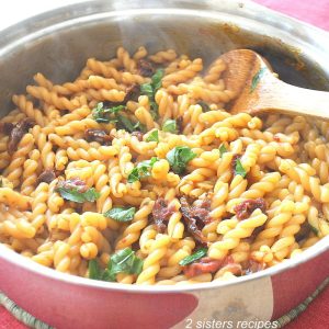 One large pot with curly pasta, fresh basil and sundried tomatoes, and a wooden spoon.