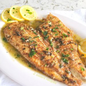 A large piece of fish cooked, and served on a white plate with lemon sauce and slices of lemon on the side.