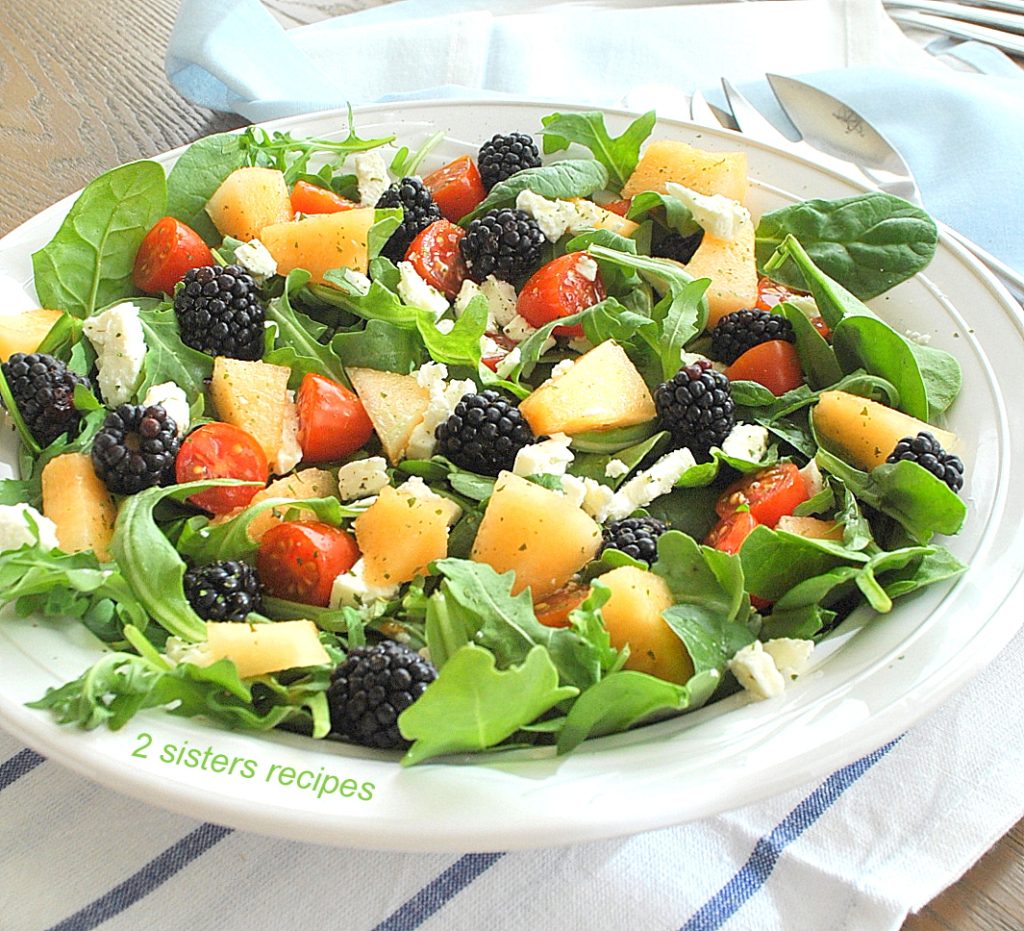 A plate filled with green salad topped with blackberries, chunks of cantaloupe and cheese.
