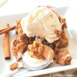 A white plate with apple crumble with scoop of vanilla ice cream on top.
