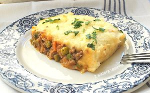 A blue and white plate with a serving of casserole with meat and peas and mashed potatoes on top.