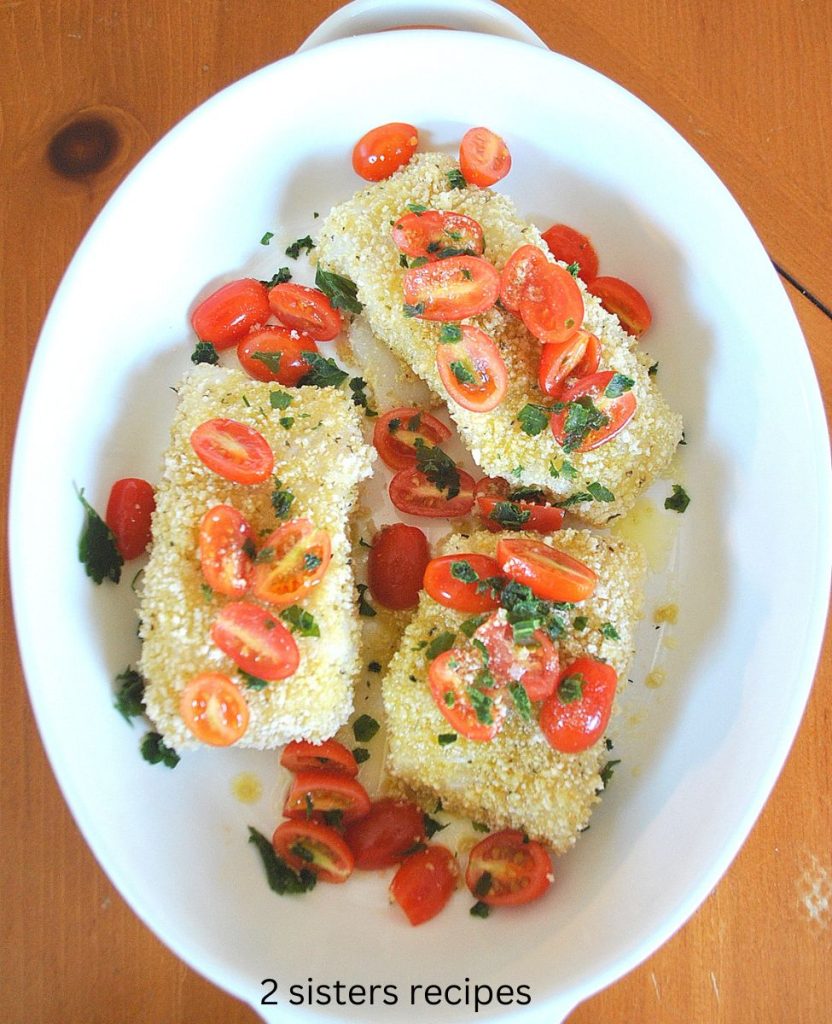 Raw pieces of cod covered with panko, fresh cherry tomatoes and parsley set in a white baking dish.