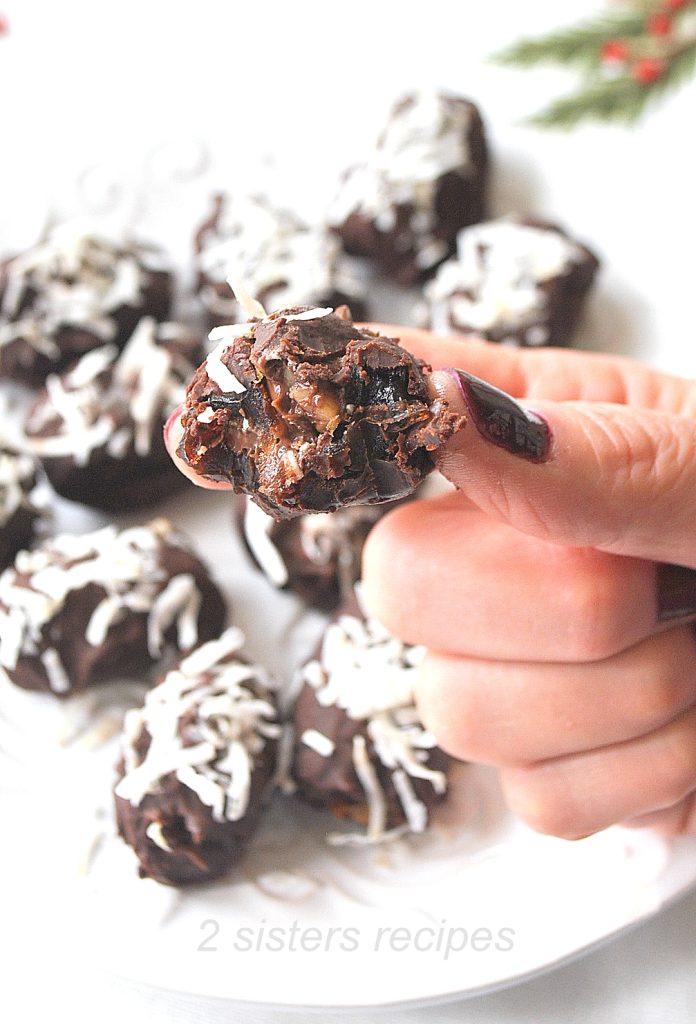 A hand holding a bite into a chocolate covered date . by 2sistersrecipes.com