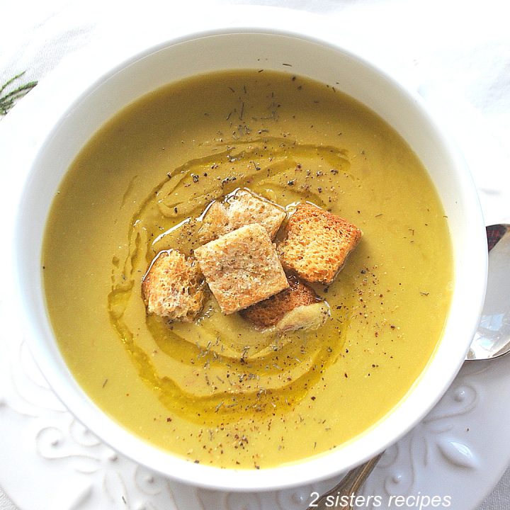 A white bowl of creamy green colored soup with croutons on top. by 2sistersrecipes.com