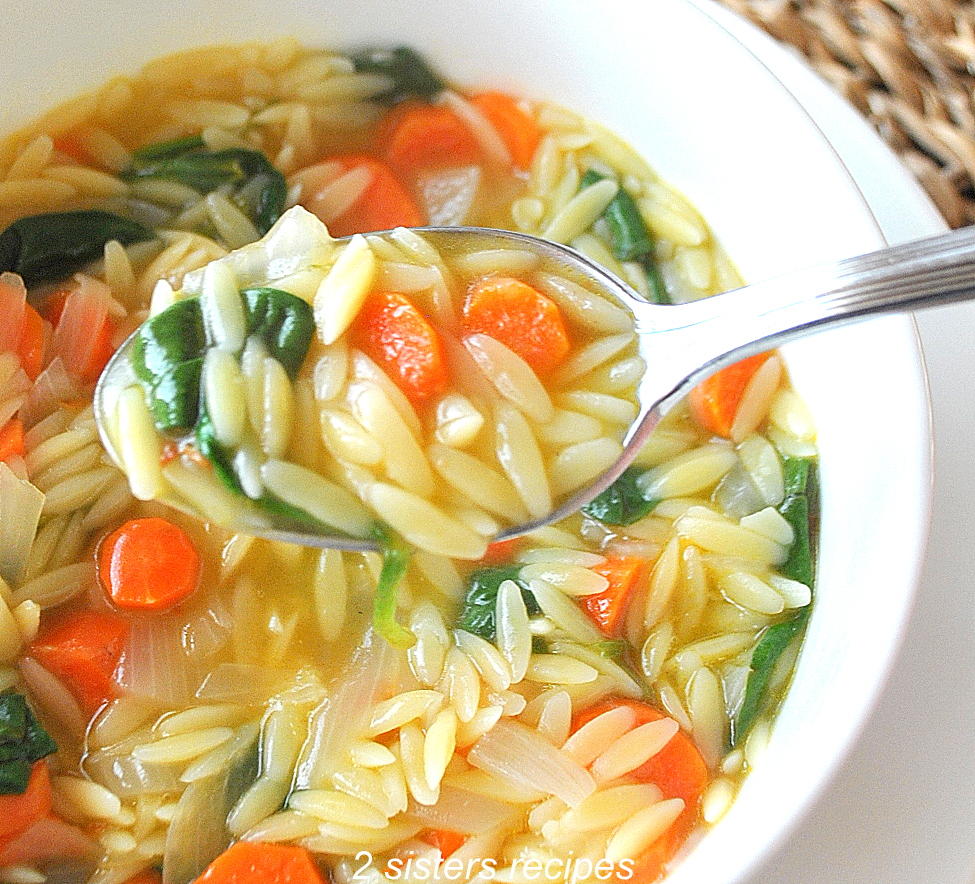 A closeup photo of spoonful of orzo, spinach and carrots soup over a white bowl. by 2sistersrecipes.com