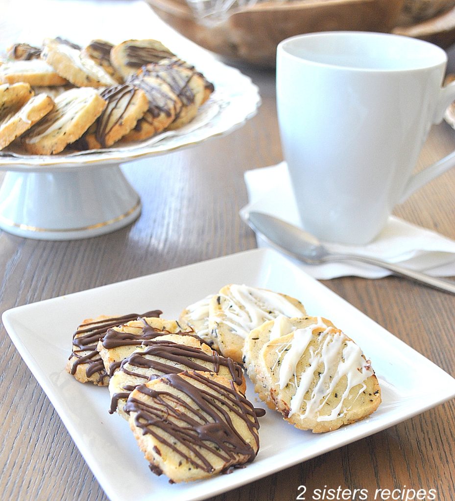 A white plate with a few cookies, along with a coffee mug and cake plate with cookies nearby. by 2sistersrecipes.com
