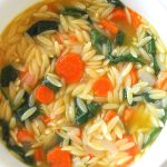 A white soup bowl filled with soup made with orzo pasta, spinach and sliced carrots.