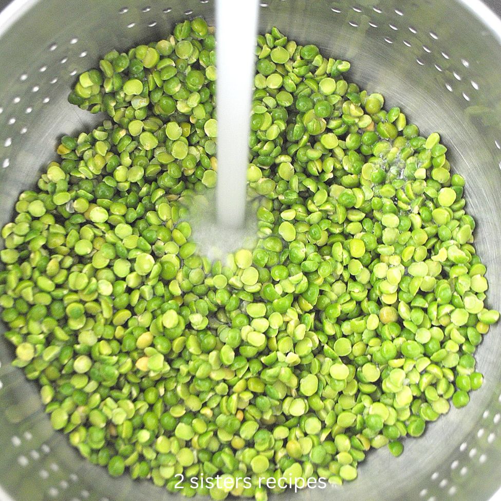 A sitainless steel colander with split green peas under running water. by 2sistersrecipes.com