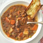 A bowl of thick soup loaded with chunks of beef, potatoes, carrots, mushrooms and peas. with a piece of crusty bread in the side.