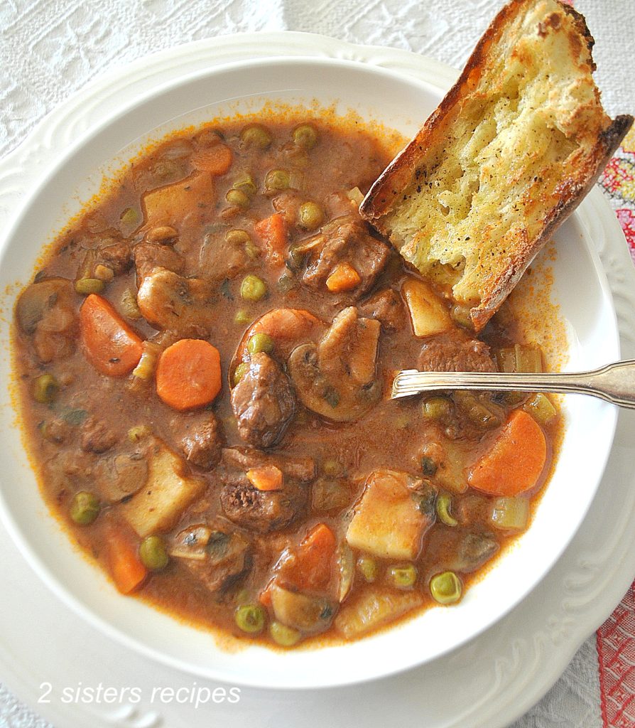 A bowl of thick soup loaded with chunks of beef, potatoes, carrots, mushrooms and peas. with a piece of crusty bread in the side. 