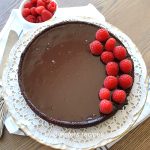A Chocolate Cake with fresh raspberries on top set on a white cake plate.