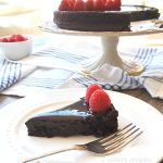 A slice of chocolate cake topped wit h chocolate ganache, and fresh raspberries served on a white plate with a fork.