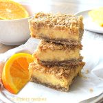 Three Orange crumb bars are piled and set on a white plate with a slice of orange on the side.