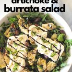 A white salad bowl filled with fresh arugula, artichoke hearts, olives and burrata cheese on top.