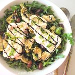 A white salad bowl filled with fresh arugula topped with olive, artichoke hearts and burrata cheese, drizzled with balsamic vinegar.