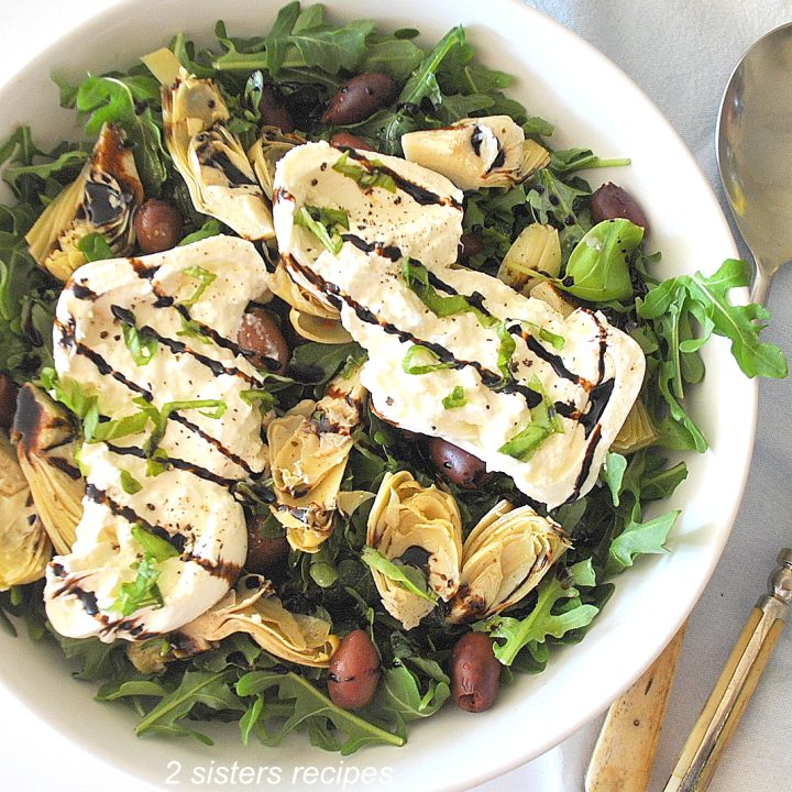 A white salad bowl filled with fresh arugula topped with olive, artichoke hearts and burrata cheese, drizzled with balsamic vinegar.