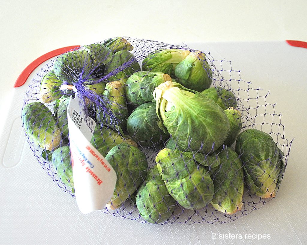 A net filled with Brussels Sprouts on a white cutting board. by 2sistersrecipes.com