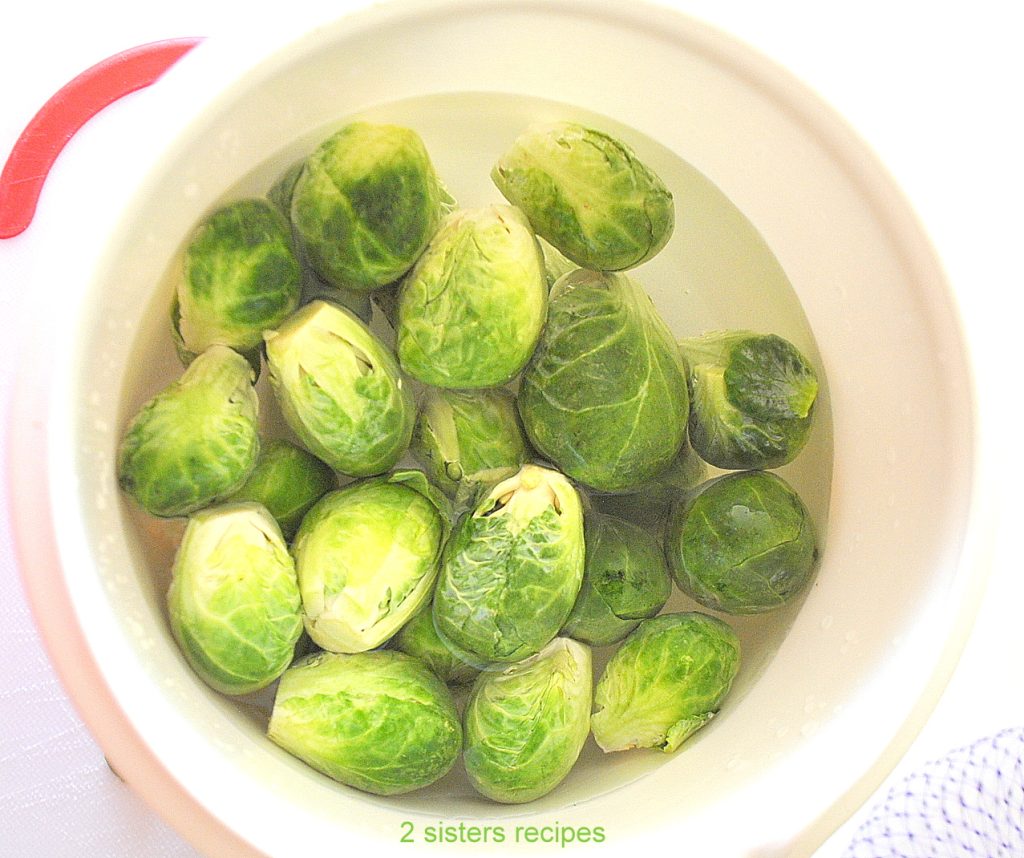 Fresh Brussels sprouts submerged in water in a white bowl.  by 2sistersrecipes.com