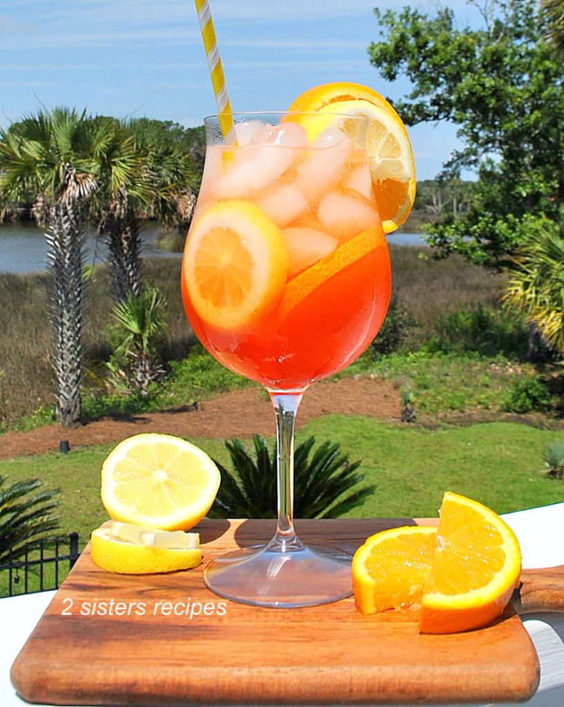 A wine glass with an orange colored hue drink with slices of oranges and lemons on a wood board.