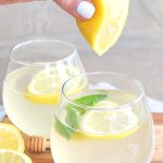 A hand squeezing a lemon into a glass of water with lemon and mint.