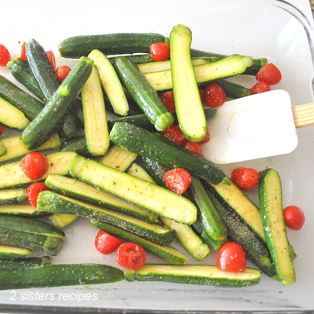 Mini zucchini and cherry tomatoes are tossed with a white spatula in a baking dish. by 2sistersrecipes.com
