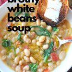 A white bowl filled with white bean soup, with spinach and a toasted bread with butter on it on the side.