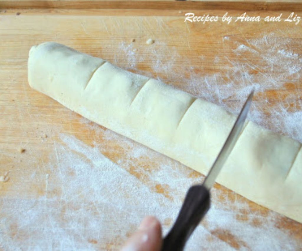 an unbaked cinnamon roll on a cutting board with knife markings on it to slice. by 2sistersrecipes.com
