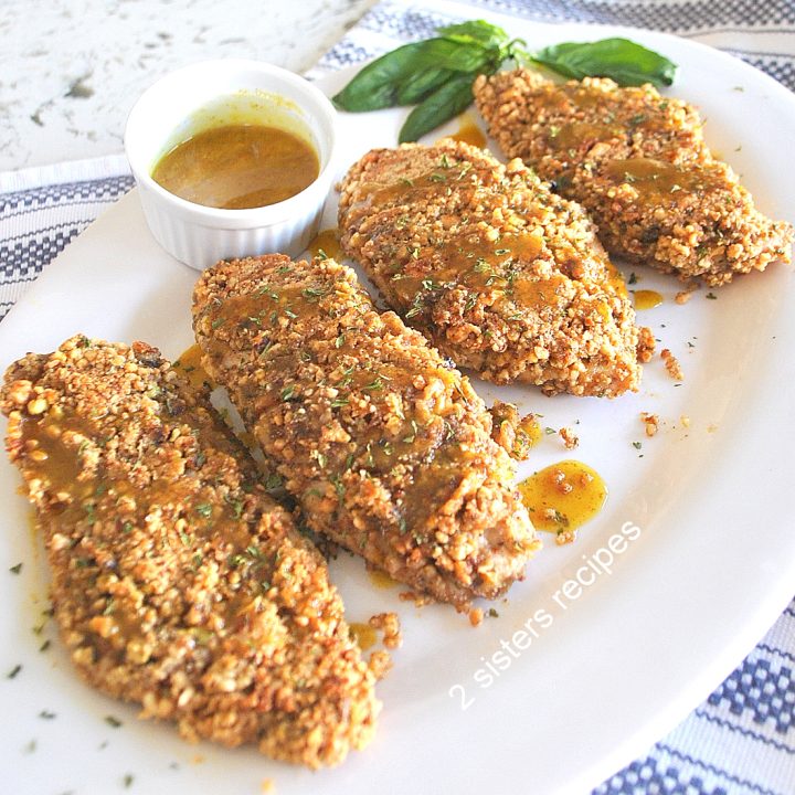 Four baked chicken cutlets coated with crushed nuts and served on a white platter with a sauce on the side.
