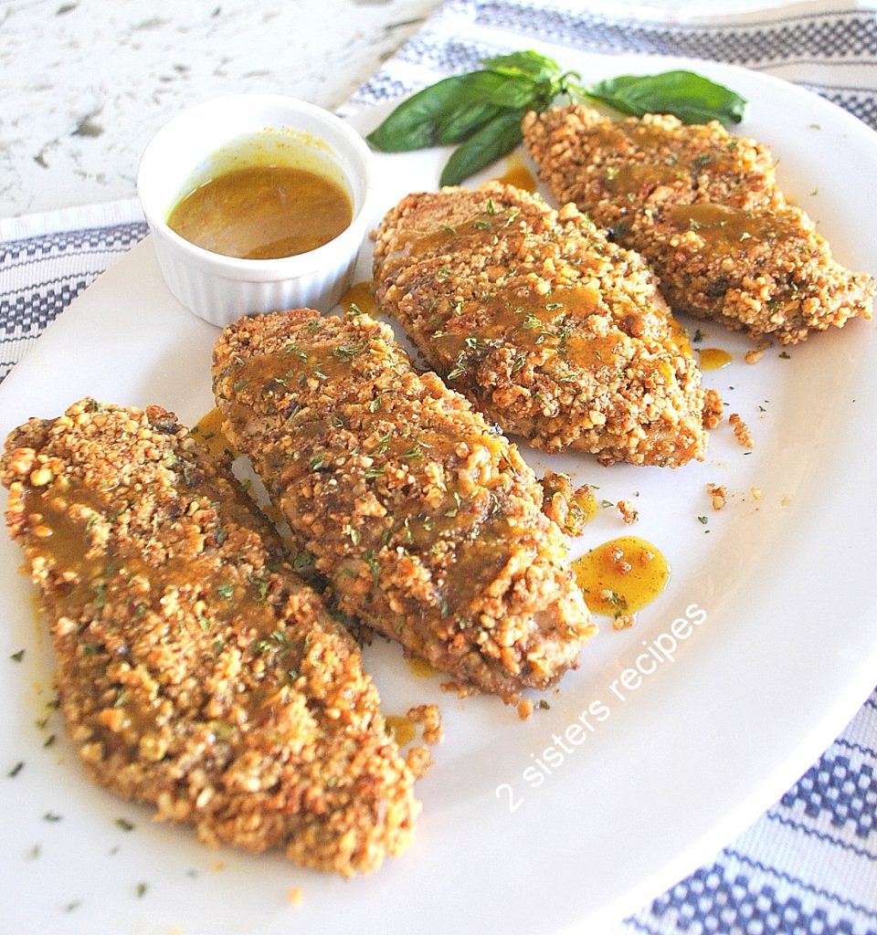 Four baked chicken cutlets coated with crushed nuts and served on a white platter with a sauce on the side.