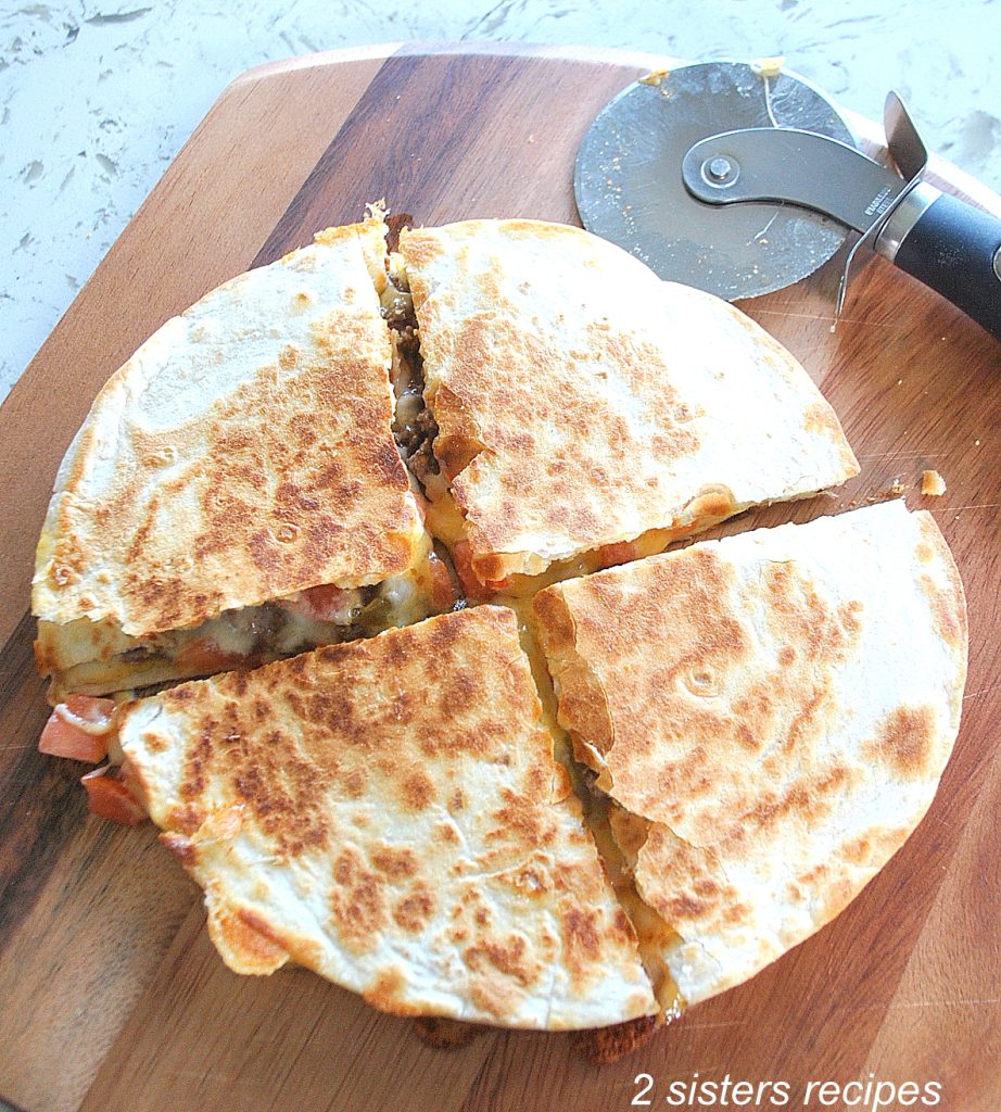 A loaded beef quesadilla is sliced into quarters on a wooden cutting board. by 2sistersrecipes.com