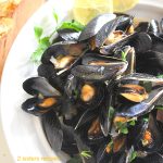 Steamed Mussels in a flavorful garlic fennel broth served in a round white bowl with lemon slices on the side.