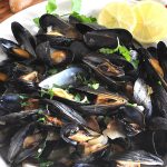 Steamed Mussels served in a white bowl with some lemons on the side.