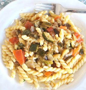A white plate filled with curly pasta and chopped vegetables with a fork resting on the plate.
