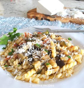 A white plate filled with curly pasta and veggies and grated cheese on top.